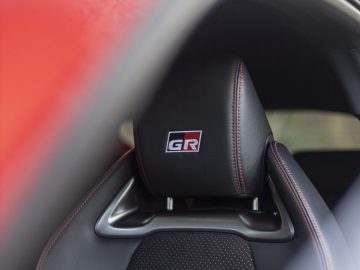 Close-up van de hoofdsteun van een autostoel met het 'GR'-logo, zwart leer en rode stiksels. Een deel van het rode exterieur van de Toyota GR Yaris is ook zichtbaar in het frame.