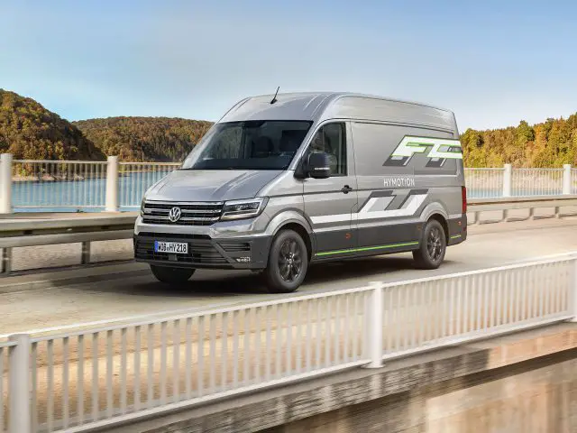 Een zilveren Volkswagen Crafter FC waterstofauto rijdt op een brug met op de achtergrond een schilderachtig uitzicht op bomen en water.