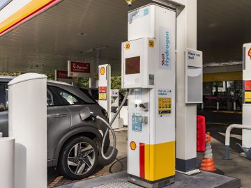 Een waterstofbrandstofpomp bij een Shell waterstofstation staat naast een auto die op de pomp is aangesloten. Zichtbare bewegwijzering geeft "Shell Hydrogen" en "V-Power" aan.