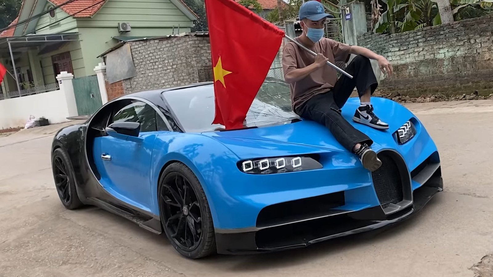 Een persoon met een pet en een masker op zit op de motorkap van een blauwe Bugatti-sportwagen met een grote rode vlag met een gele ster, geparkeerd in een woonstraat.