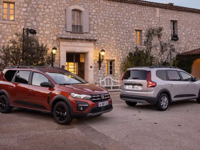 Twee moderne SUV's, waaronder een strakke Dacia Jogger, staan geparkeerd voor een rustiek stenen gebouw met verlichte lantaarns in de schemering. De ene SUV is rood en de andere is zilver.