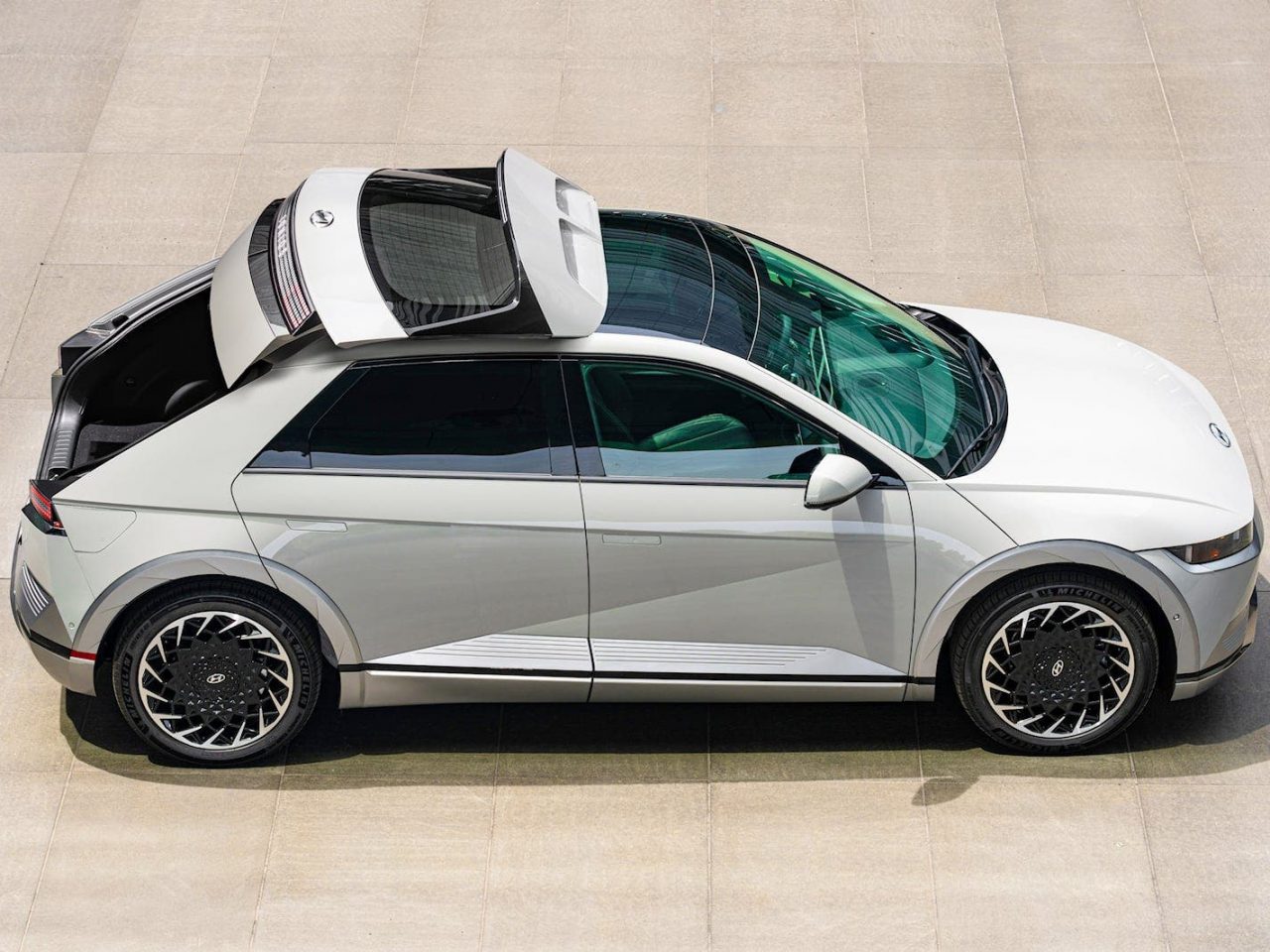 Een witte compacte Hyundai-auto met het kofferluik open, gezien vanuit een bovenhoek op een beige betonnen oppervlak. De auto beschikt over moderne designkenmerken en zwarte velgen met cirkelvormig patroon.