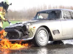 Een brandweerman in uitrusting gebruikt een slang om de vlammen te doven die uit de voorkant van een beschadigde Maserati 3500 GTI op een natte weg komen.