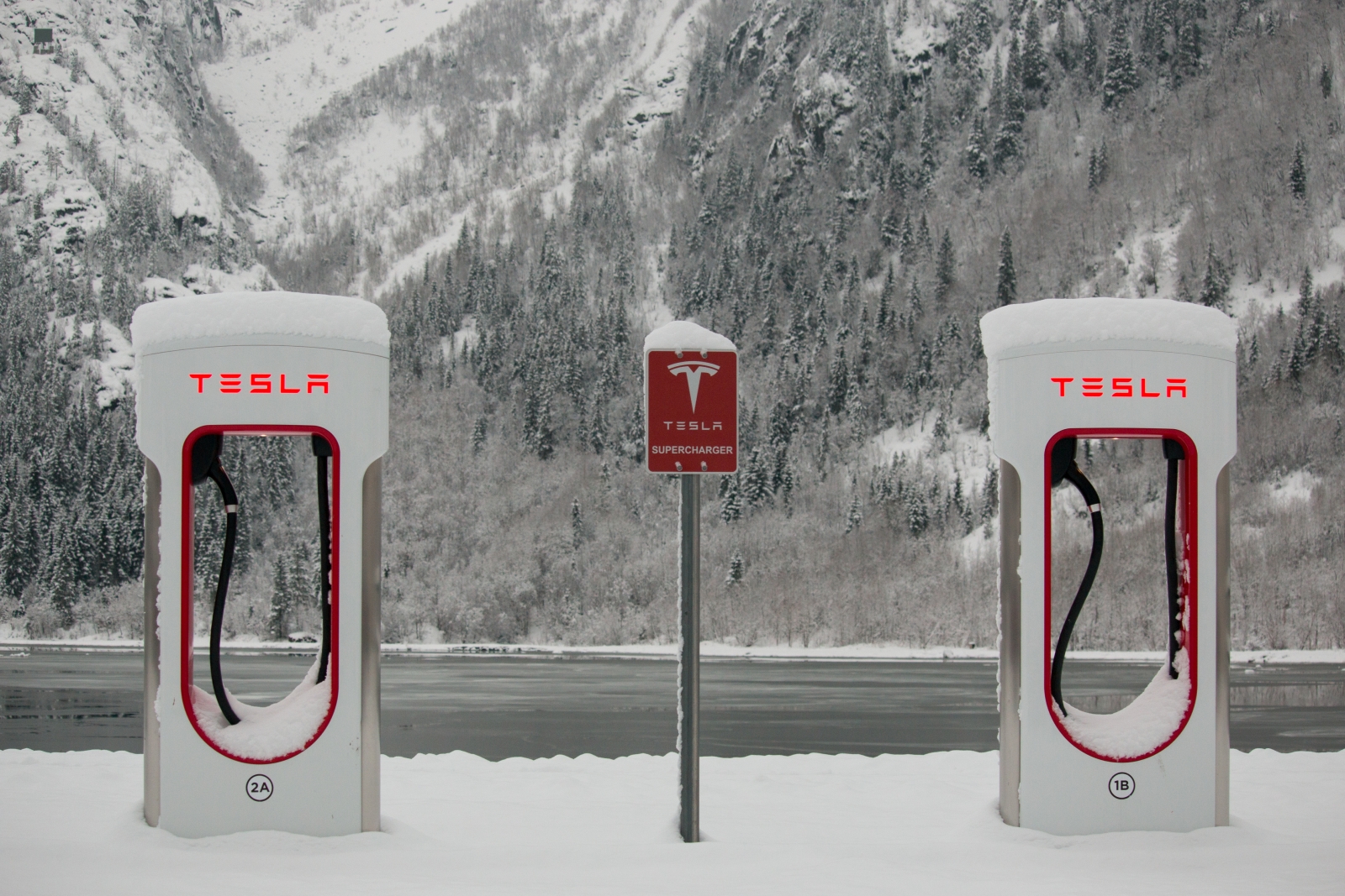 Twee met sneeuw bedekte Tesla Supercharger-stations staan tegen een adembenemende achtergrond van besneeuwde bergen en bomen, perfect voor uw wintersportavonturen.