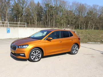 Een metallic oranje Skoda Fabia hatchback staat geparkeerd op een grindoppervlak met een bosgebied op de achtergrond en aan de linkerkant is een bord met een parkeerplaats zichtbaar.