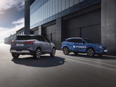 Twee auto's geparkeerd voor een modern gebouw op een zonnige dag, met de auto aan de rechterkant trots gehuld in het Qashqai e-POWER-logo.