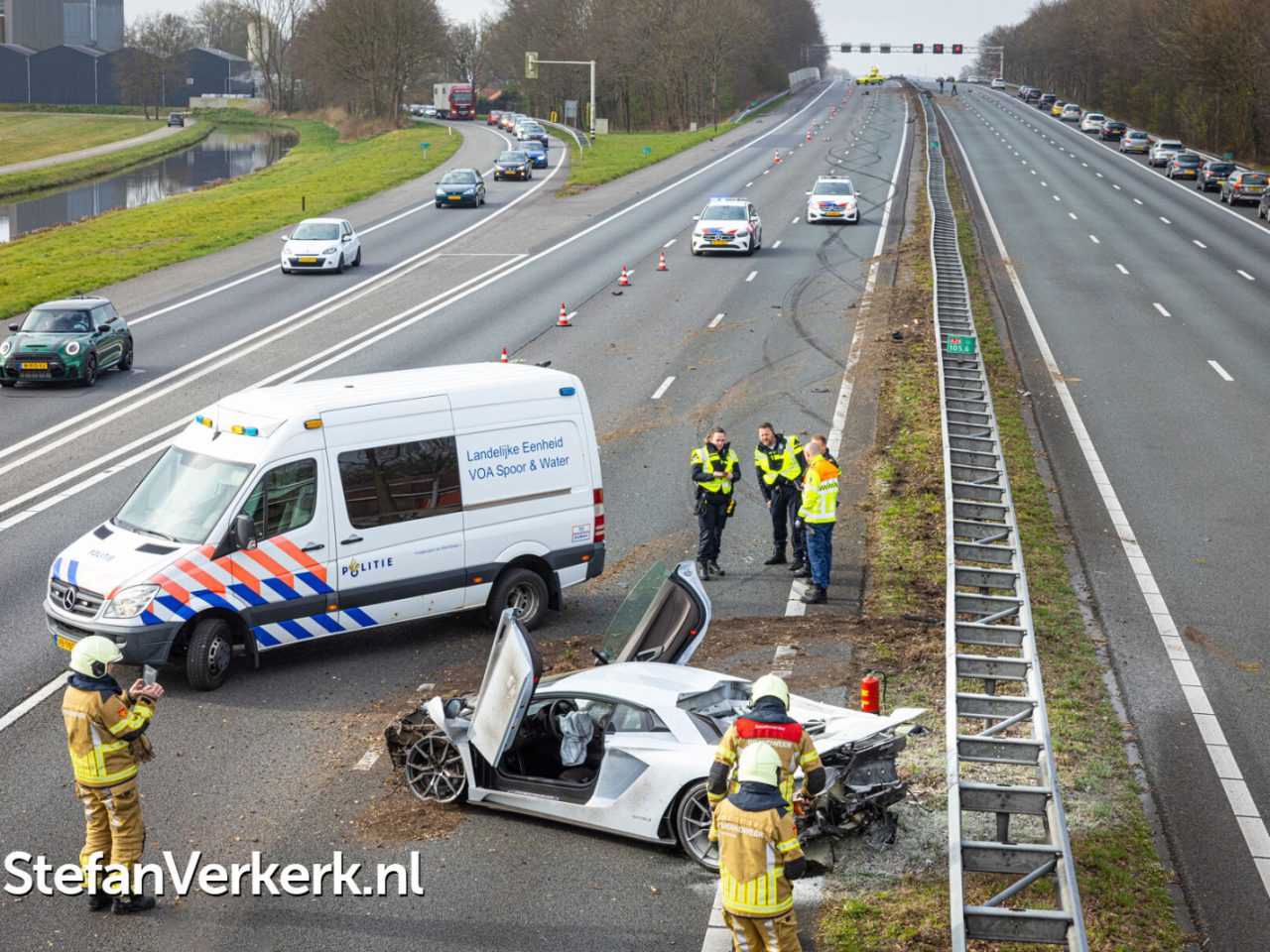 Door een Lamborghini-ongeluk is een witte sportwagen zwaar beschadigd geraakt op de middenberm van een snelweg met meerdere rijstroken. Politieagenten en brandweerlieden zijn ter plaatse en er staat een hulpvoertuig in de buurt geparkeerd. Het verkeer wordt aan de overkant tegengehouden.