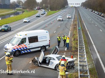 Door een Lamborghini-ongeluk is een witte sportwagen zwaar beschadigd geraakt op de middenberm van een snelweg met meerdere rijstroken. Politieagenten en brandweerlieden zijn ter plaatse en er staat een hulpvoertuig in de buurt geparkeerd. Het verkeer wordt aan de overkant tegengehouden.
