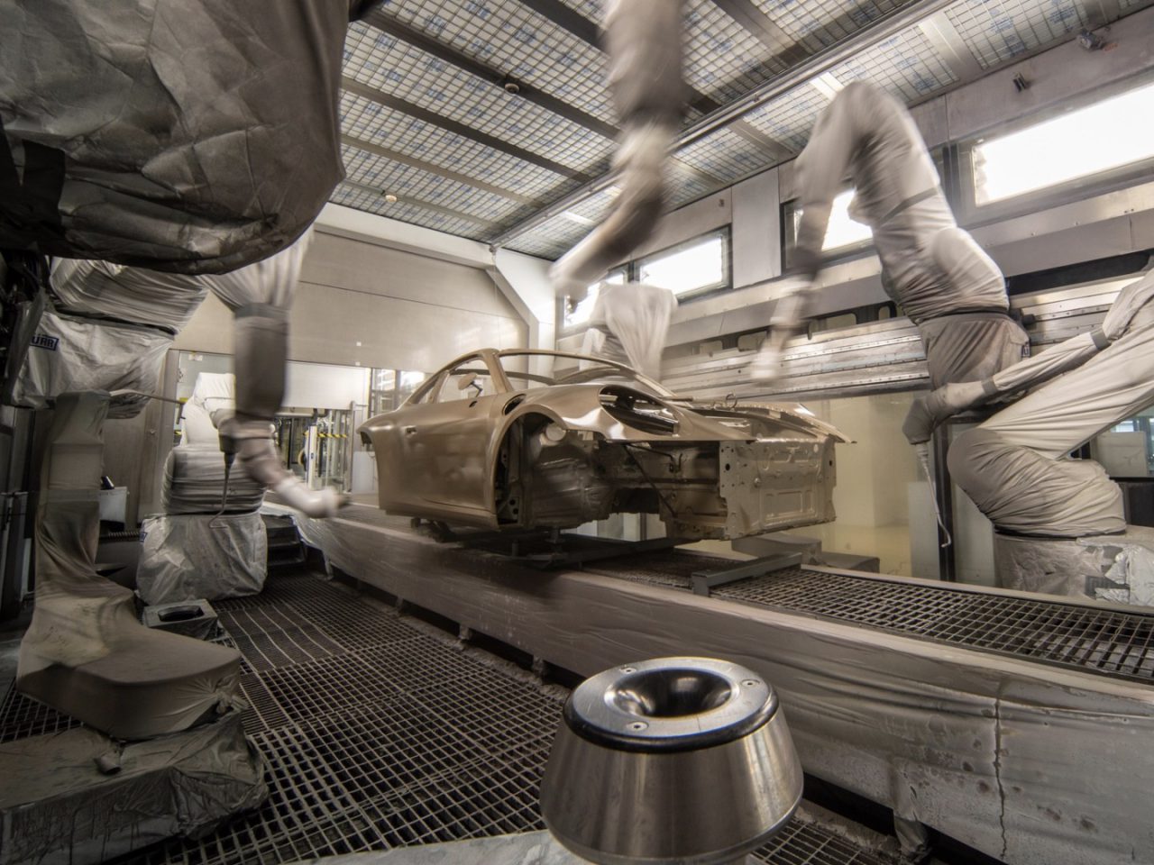 Geautomatiseerde robotarmen brengen nauwgezet Porsche-verf aan om een carrosserie te bemonsteren in een industriële spuitcabine. De auto hangt aan een transportsysteem en de kamer is gevuld met veiligheidsapparatuur en geavanceerde ventilatiesystemen.