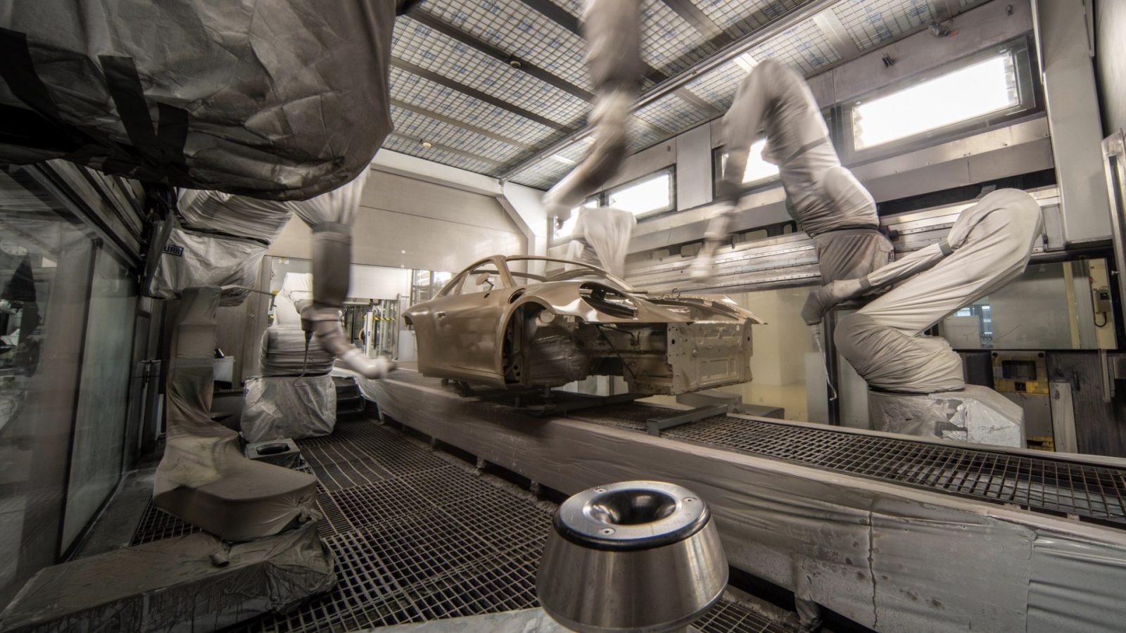 Geautomatiseerde robotarmen brengen nauwgezet Porsche-verf aan om een carrosserie te bemonsteren in een industriële spuitcabine. De auto hangt aan een transportsysteem en de kamer is gevuld met veiligheidsapparatuur en geavanceerde ventilatiesystemen.