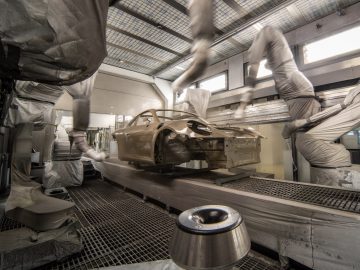 Geautomatiseerde robotarmen brengen nauwgezet Porsche-verf aan om een carrosserie te bemonsteren in een industriële spuitcabine. De auto hangt aan een transportsysteem en de kamer is gevuld met veiligheidsapparatuur en geavanceerde ventilatiesystemen.