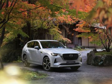 Een witte Mazda CX-60 SUV staat geparkeerd op een grindpad onder bomen met herfstbladeren, grenzend aan een traditioneel ogend gebouw.