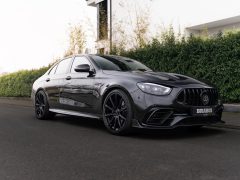 Een zwarte luxe sedan, met trots een ‘BRABUS’-embleem op de grille, staat geparkeerd op straat. Hij heeft een strak, aerodynamisch ontwerp met zwarte velgen. De achtergrond toont weelderig groen en een modern gebouw.