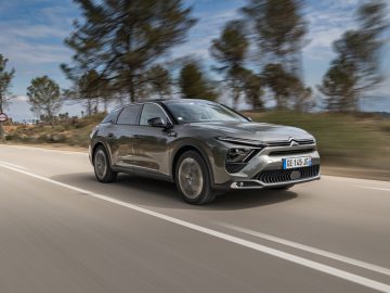 Een slanke zilveren Citroën C5 X glijdt over de weg, met vage bomen op de achtergrond onder een blauwe lucht bezaaid met verspreide wolken.