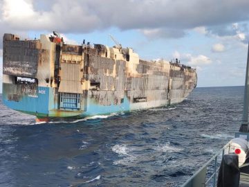 Een verbrand en beschadigd vrachtschip drijft in de oceaan, wat doet denken aan het recente incident waarbij een schip auto's gezonken ontmoette. Een ander schip met reddingsboeien is gedeeltelijk zichtbaar op de voorgrond. De lucht is gedeeltelijk bewolkt.