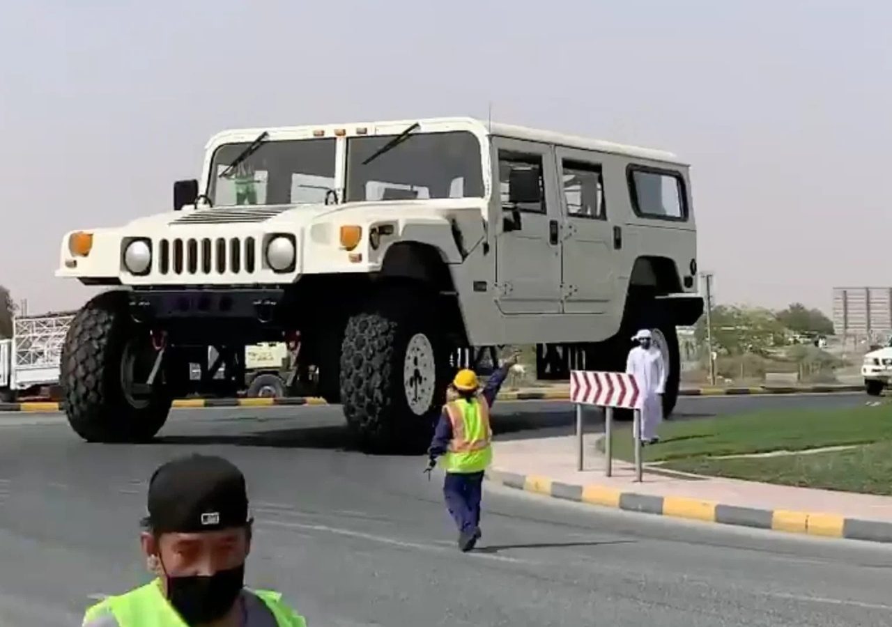 Een grote Hummer met extra grote banden wordt over de weg gereden. Bij het verhoogde voertuig worden twee bouwvakkers in veiligheidsvesten en een man in traditionele kledij gezien.