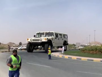Een grote witte Hummer wordt over de weg gereden met hulp van twee mensen met veiligheidsvesten die het verkeer regelen.