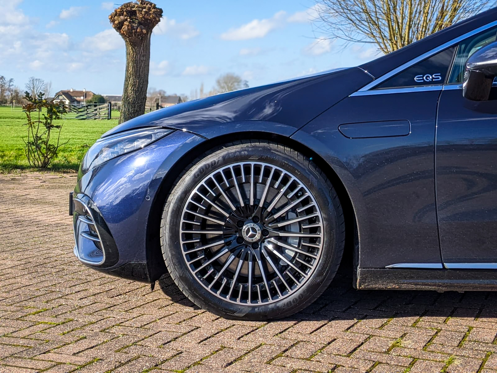Close-up zijaanzicht van het voorwiel, de band en een deel van de motorkap van een donkerblauwe Mercedes EQS, geparkeerd op een bakstenen stoep met een boom en een groen veld op de achtergrond.