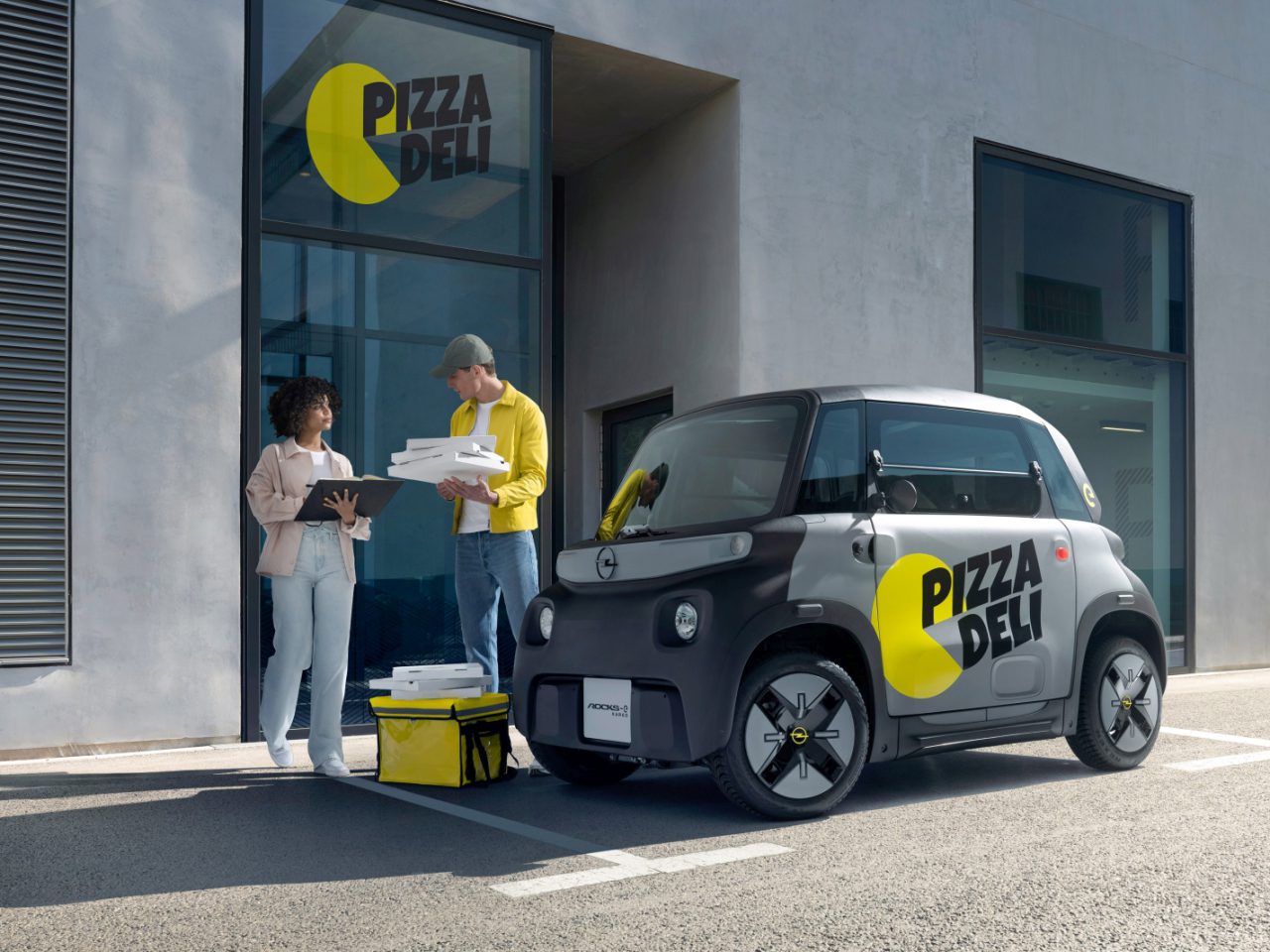 Twee personen staan buiten een gebouw met de bewegwijzering "Pizza Deli". De een houdt pizzadozen vast, terwijl de ander op een klembord kijkt. Een compacte Opel Rocks-e KARGO, met het opschrift ‘Pizza Deli’, staat vlakbij geparkeerd.