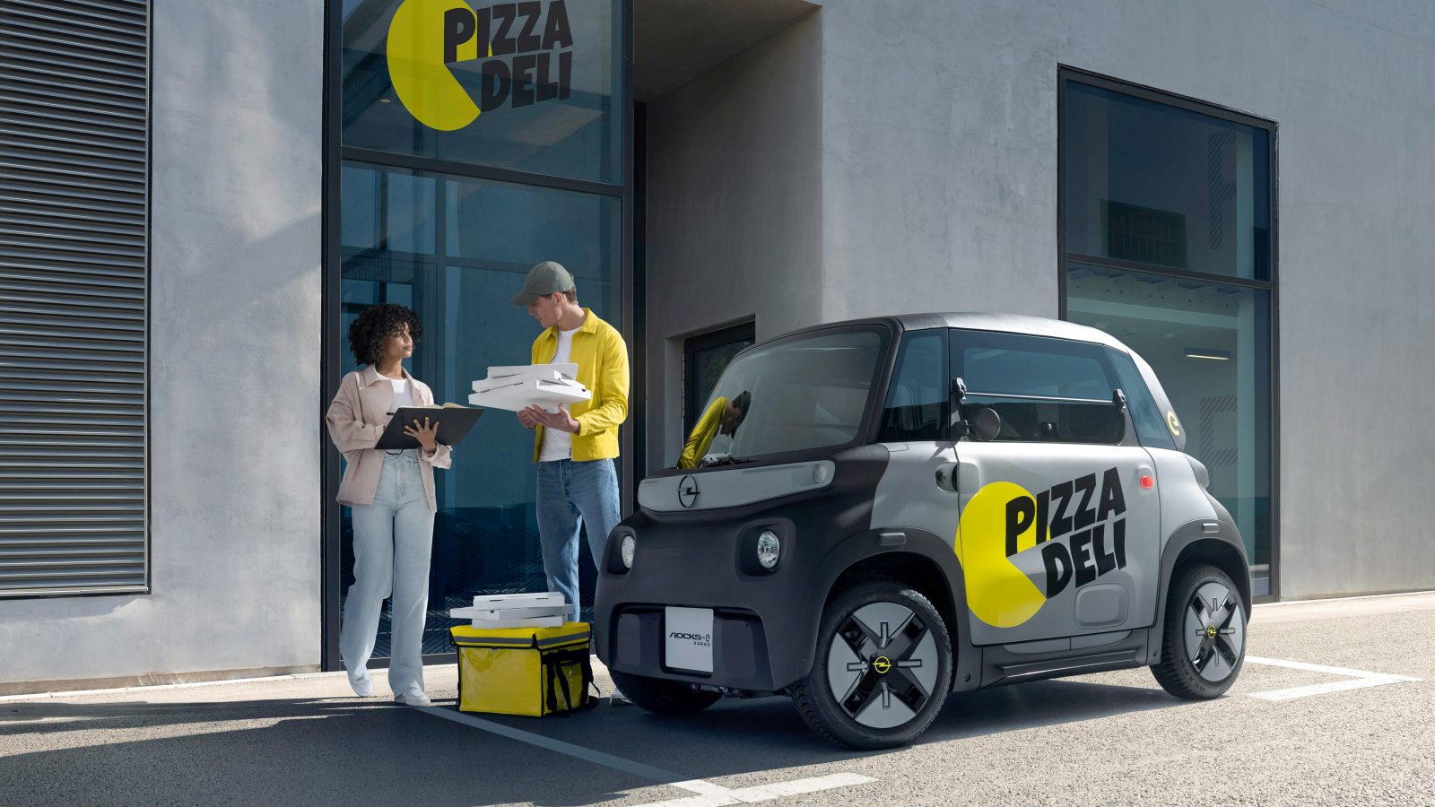 Twee personen staan buiten een gebouw met de bewegwijzering "Pizza Deli". De een houdt pizzadozen vast, terwijl de ander op een klembord kijkt. Een compacte Opel Rocks-e KARGO, met het opschrift ‘Pizza Deli’, staat vlakbij geparkeerd.