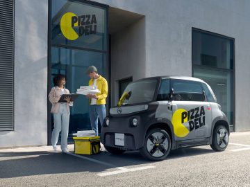 Twee personen staan buiten een gebouw met de bewegwijzering "Pizza Deli". De een houdt pizzadozen vast, terwijl de ander op een klembord kijkt. Een compacte Opel Rocks-e KARGO, met het opschrift ‘Pizza Deli’, staat vlakbij geparkeerd.