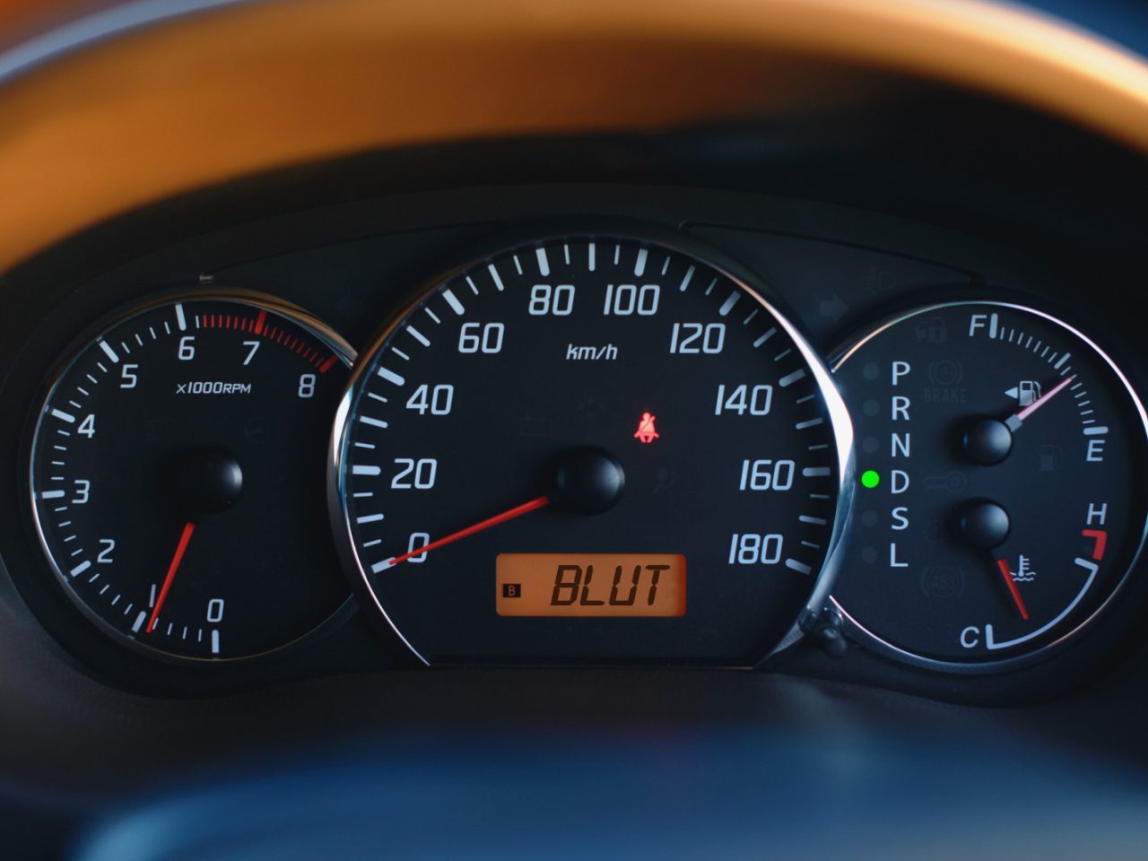 Close-up van een autodashboard met snelheidsmeter, toerenteller, brandstofmeter en versnellingsindicator. De snelheidsmeter geeft 120 km/u aan en op het digitale display staat 'BLUT', terwijl er wordt nagedacht over de impact op uw kilometervergoeding.