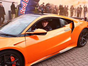 Te midden van een achtergrond van oranje rook en een prominent Red Bull-spandoek zit een persoon zelfverzekerd op de bestuurdersstoel van een oranje sportwagen, die doet denken aan die van Max Verstappen. Toeschouwers en fotografen leggen gretig het moment vast.