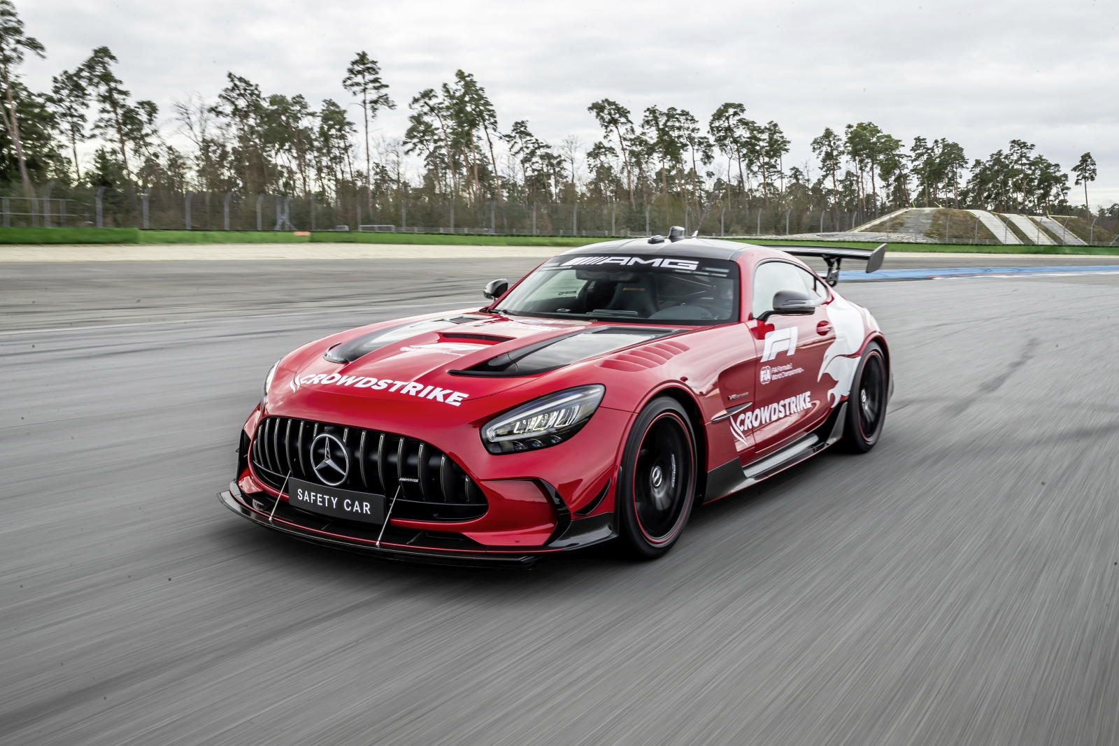 Een rode Mercedes-AMG safety car, versierd met 'Crowdstrike', rijdt op een racecircuit omgeven door bomen. Het gestroomlijnde voertuig is een vaste waarde tijdens Formule 1-evenementen en zorgt voor soepele en veilige raceomstandigheden.