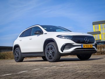 Een witte Mercedes-Benz 350 4MATIC SUV staat geparkeerd op een verhard terrein onder een helderblauwe hemel. Op het kenteken aan de voorkant staat "N-902-ZR." Op de achtergrond is een geel en zwart gebouw zichtbaar.