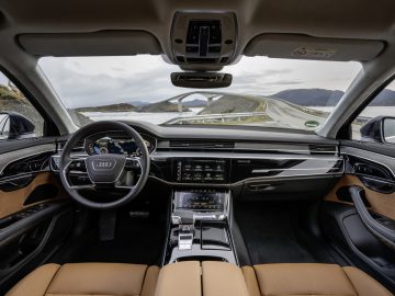 Binnenaanzicht van een modern Auto Koninklijk Huis-voertuig, met het dashboard, het stuur, de middenconsole met verschillende bedieningselementen en schermen, en een schilderachtige kustweg die zich door de voorruit uitstrekt.