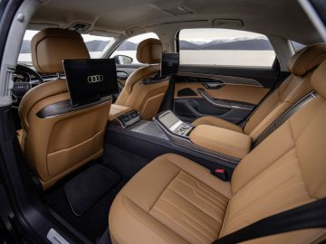 Het interieur van een luxe auto van Auto Koninklijk Huis is voorzien van bruin lederen stoelen, donkerhouten bekleding, twee schermen aan de achterkant van de voorstoelen en een middenconsole. Door de ramen kunt u genieten van een schilderachtig uitzicht op bergen en water.