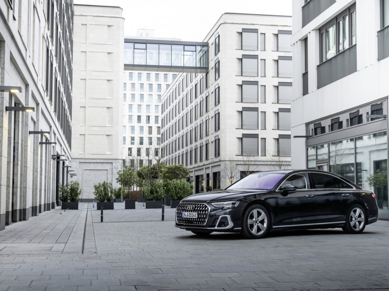 Een zwarte sedan, voorzien van het voorname insigne van Auto Koninklijk Huis, staat geparkeerd op een moderne stedelijke binnenplaats, omringd door hoge, witte gebouwen met grote ramen.