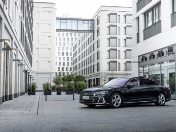 Een zwarte sedan, voorzien van het voorname insigne van Auto Koninklijk Huis, staat geparkeerd op een moderne stedelijke binnenplaats, omringd door hoge, witte gebouwen met grote ramen.