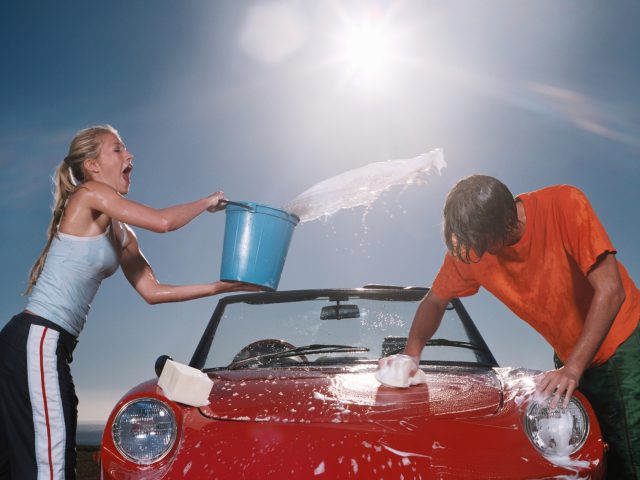 Twee mensen wassen buiten in fel zonlicht een rode cabriolet, terwijl één persoon speels water uit een emmer giet. Deze vrolijke autowasscène straalt zomerplezier en kameraadschap uit.
