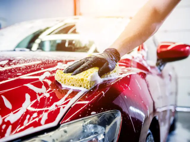 Een persoon die een zwarte handschoen draagt, wast een rode auto met een gele spons, waardoor tijdens een autowassessie zeepbellen op het oppervlak van de auto ontstaan.