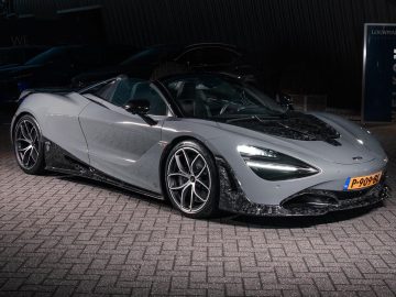 Een strakke, grijze sportwagen met cabriokap staat geparkeerd op een bakstenen oppervlak, verlicht door zachte verlichting. Het strakke koetswerkdesign en de lichtmetalen velgen zijn duidelijk zichtbaar, net als de precisie en kracht die Rico Verhoeven belichaamt.