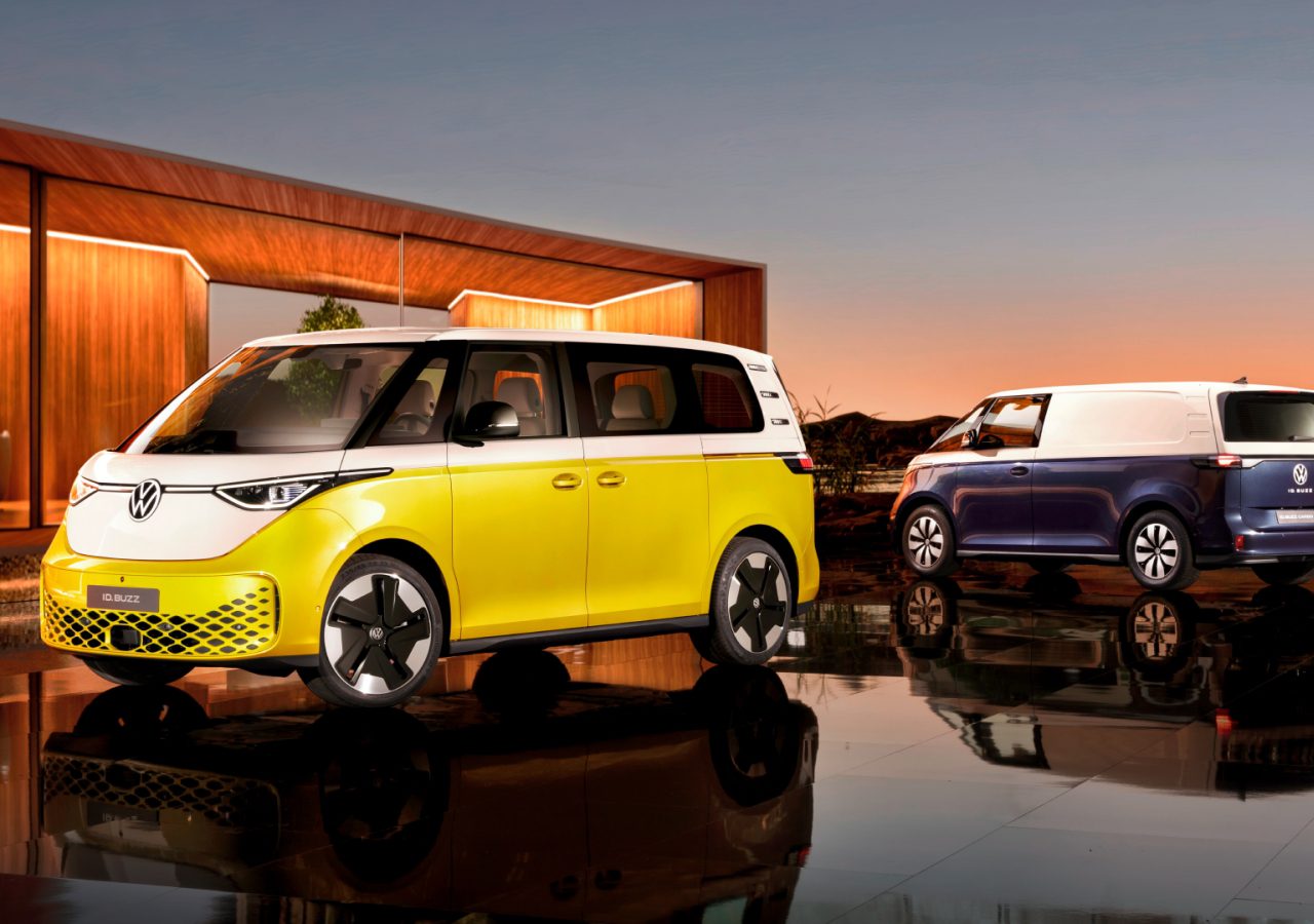 Twee moderne Volkswagenbusjes, één geel-wit en één blauw-wit, staan geparkeerd op een reflecterend oppervlak met een eigentijds gebouw en een zonsondergang op de achtergrond, wat het strakke ontwerp van de Volkswagen ID laat zien. Ophef.