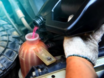 Een persoon die handschoenen draagt, giet voorzichtig roze motorkoelvloeistof, ervoor zorgend dat er geen olie in de koelvloeistof zit, vanuit een zwarte container in het koelvloeistofreservoir van de motor van een voertuig.