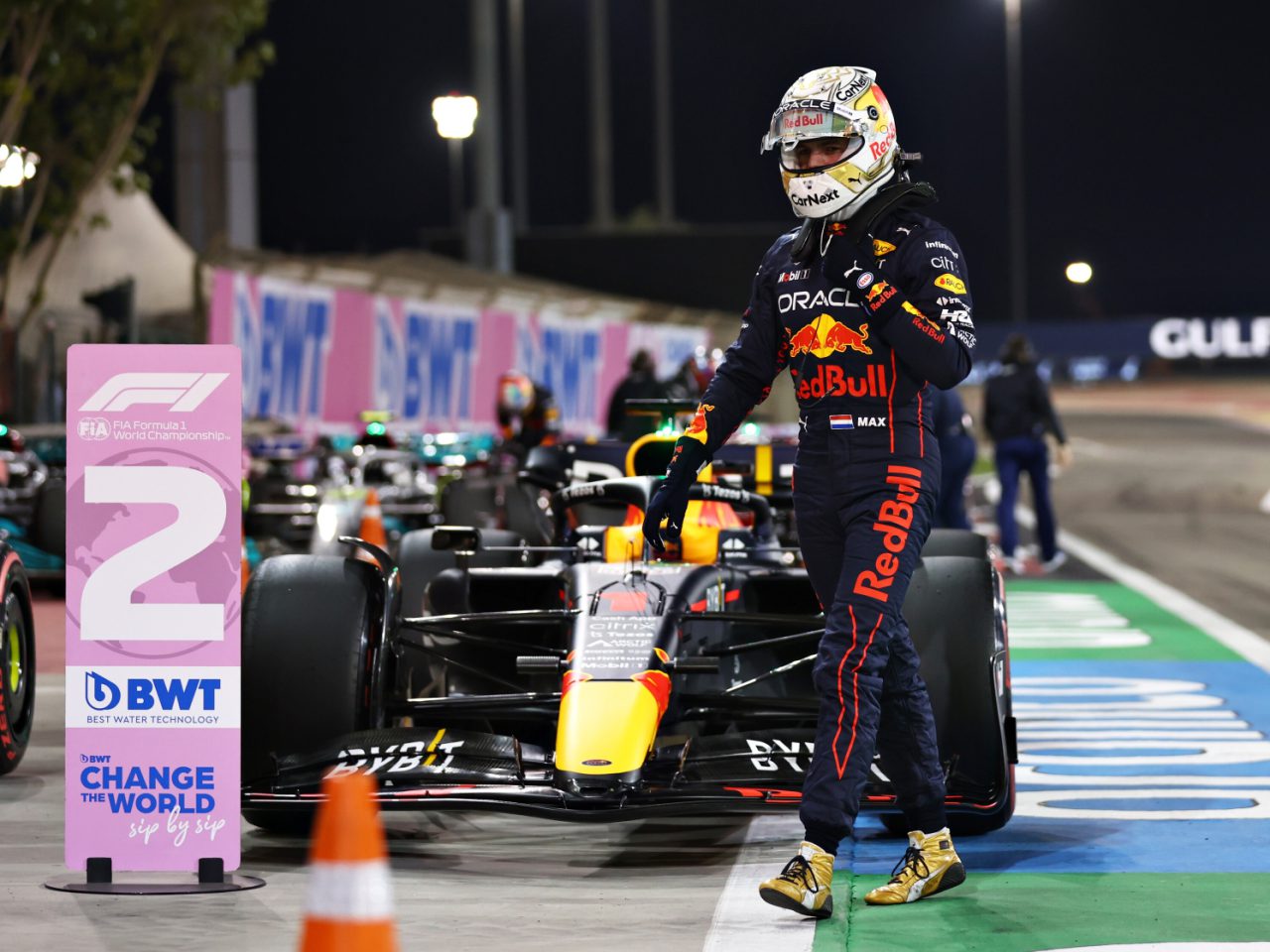 Een coureur in een Red Bull-racepak en -helm staat 's nachts naast een "2"-podiumbord op een racecircuit, met geparkeerde Formule 1-auto's en pitcrewleden op de achtergrond, waardoor fans zich afvragen: Formule 1 hoe laat?