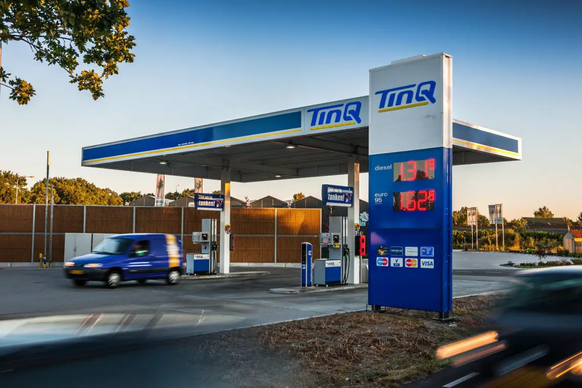 Een blauw-wit TMQ-tankstation met een digitaal bord met de prijzen voor diesel en loodvrij, perfect voor goedkope tanken. Voertuigen rijden het station in en uit, terwijl op de achtergrond bomen en gebouwen te zien zijn.