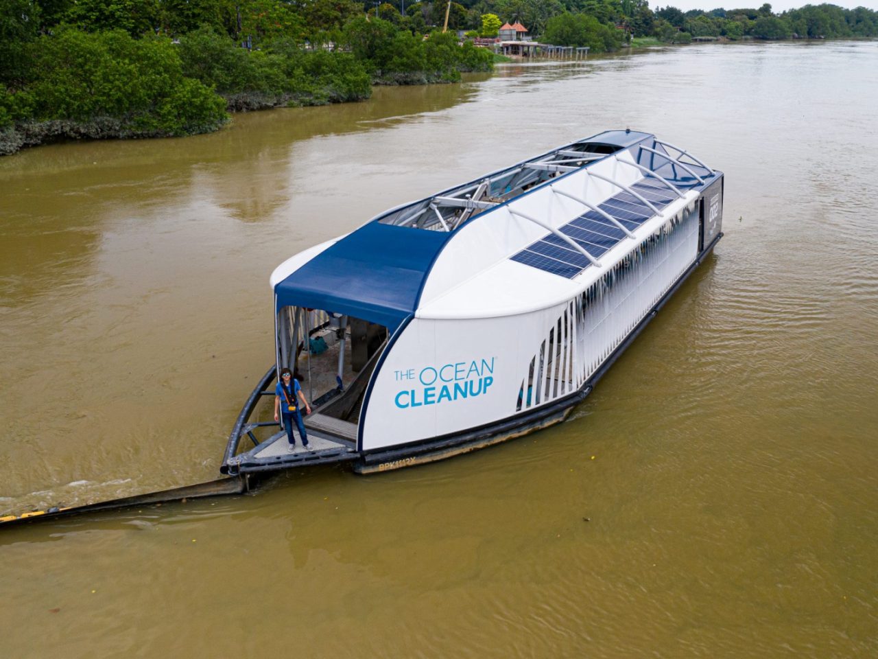 Een groot schoonmaakschip met zonnepanelen op het dak, genaamd "The Ocean Cleanup", opgericht door Boyan Slat, vaart op een rivier om afval op te halen. Er zijn twee mensen aan boord te zien terwijl bomen langs de rivieroever staan.