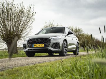 Een zilveren Audi Q5 Sportback 2022 met geel Europees kenteken rijdt op een smalle landelijke weg omzoomd met bomen en gras, onder een bewolkte hemel.