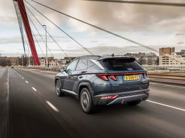 Een grijze Hyundai SUV, bekend om zijn grote kofferbak, rijdt met rode kabels over een stadsbrug, weg van de camera. Het stadsbeeld en de gebouwen zijn zichtbaar op de achtergrond onder een bewolkte hemel.