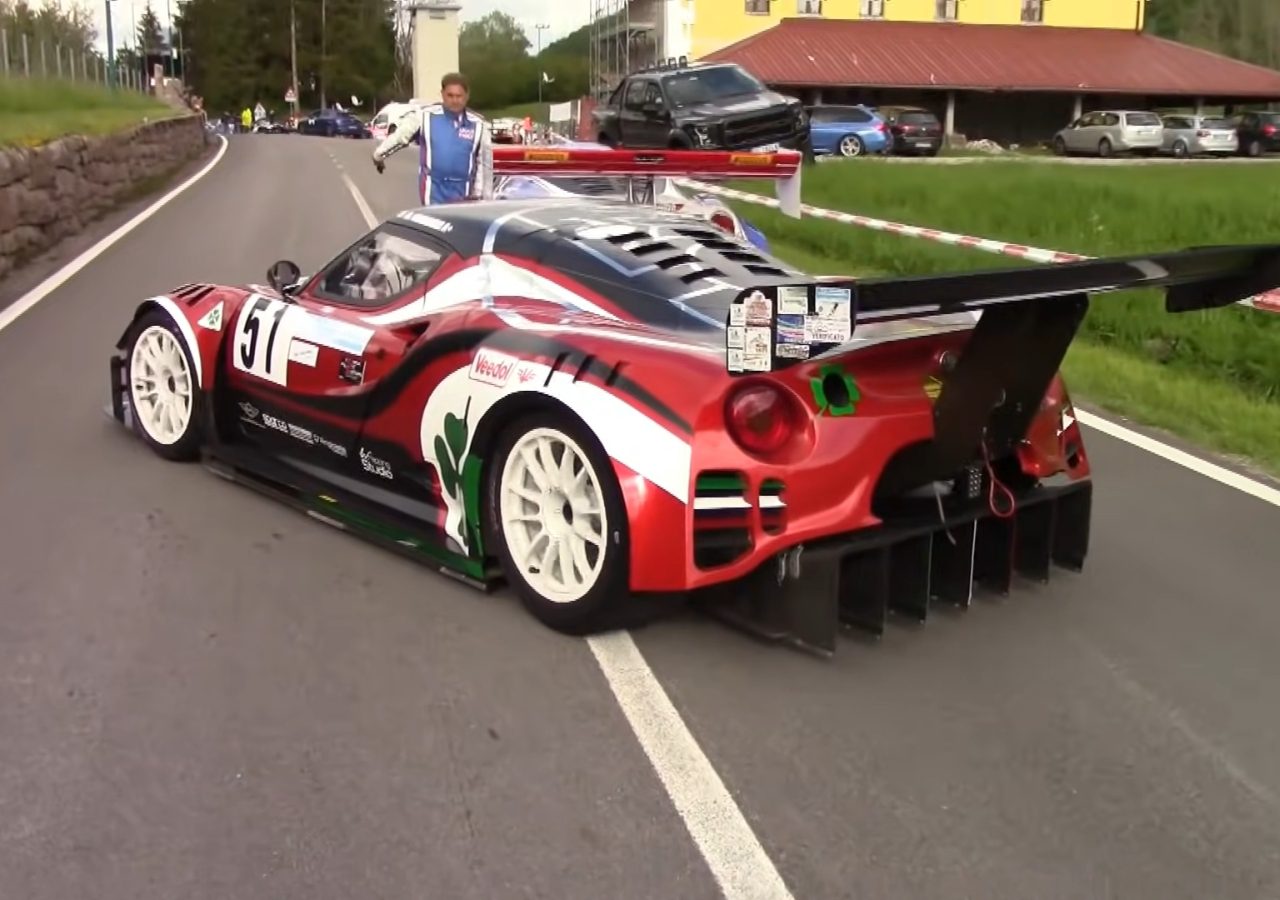 Een rood-zwarte raceauto met nummer 61, voorzien van een grote achtervleugel en witte banden, staat geparkeerd op een verharde weg nabij een groen grasveld. Met het herkenbare Formule 1-geluid op de achtergrond staat er een persoon naast de auto.
