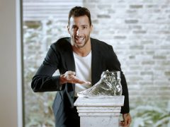 Man in een donkere blazer die een helderglazen wijnkaraf presenteert op een wit voetstuk in een kamer met een bakstenen muurachtergrond.