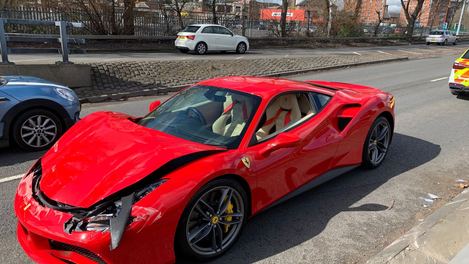 Een rode Ferrari met een beschadigde voorbumper staat langs de kant van de weg geparkeerd, mogelijk na een recente crash. Eromheen staan andere voertuigen en een bakstenen gebouw op de achtergrond.