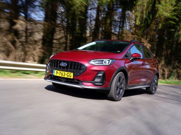 Een Nieuwe Ford Fiesta met zichtbaar kenteken rijdt op een weg met een bosrijke achtergrond.