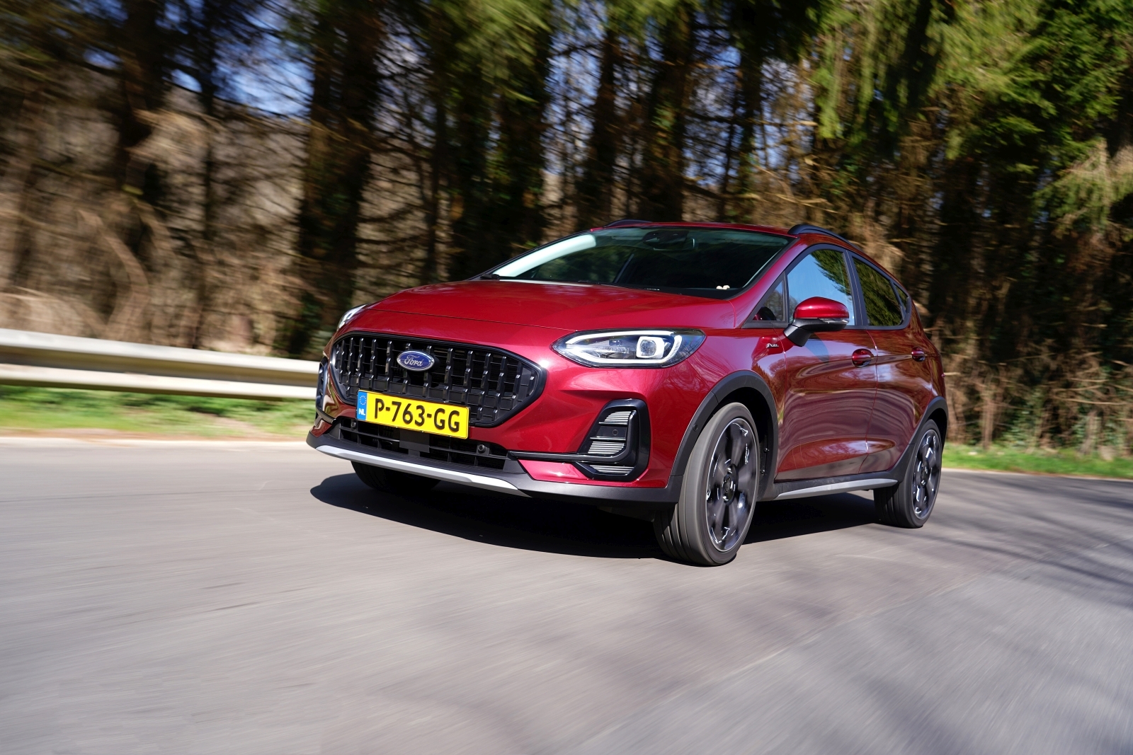 Een Nieuwe Ford Fiesta met zichtbaar kenteken rijdt op een weg met een bosrijke achtergrond.