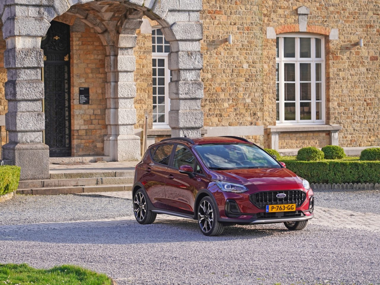 Een rode compacte Nieuwe Ford Fiesta met geel Europees kenteken staat geparkeerd voor een stenen gebouw met een gewelfde ingang en een groot raam.