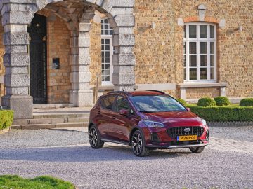 Een rode compacte Nieuwe Ford Fiesta met geel Europees kenteken staat geparkeerd voor een stenen gebouw met een gewelfde ingang en een groot raam.